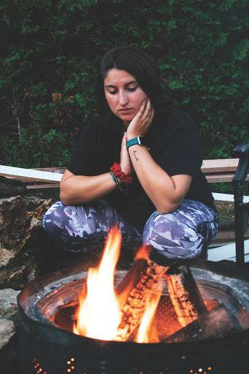 A photo of a young girl with brown hair crouching in front of a fire. She has a serious expression on her face while resting her hand on her face with her hand. 