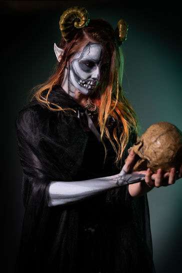 A petit woman with red hair and.a black dress holds a skull. She has horns, and the ears of a mythical creature. She stands, starting intently into the eyes of the skull.